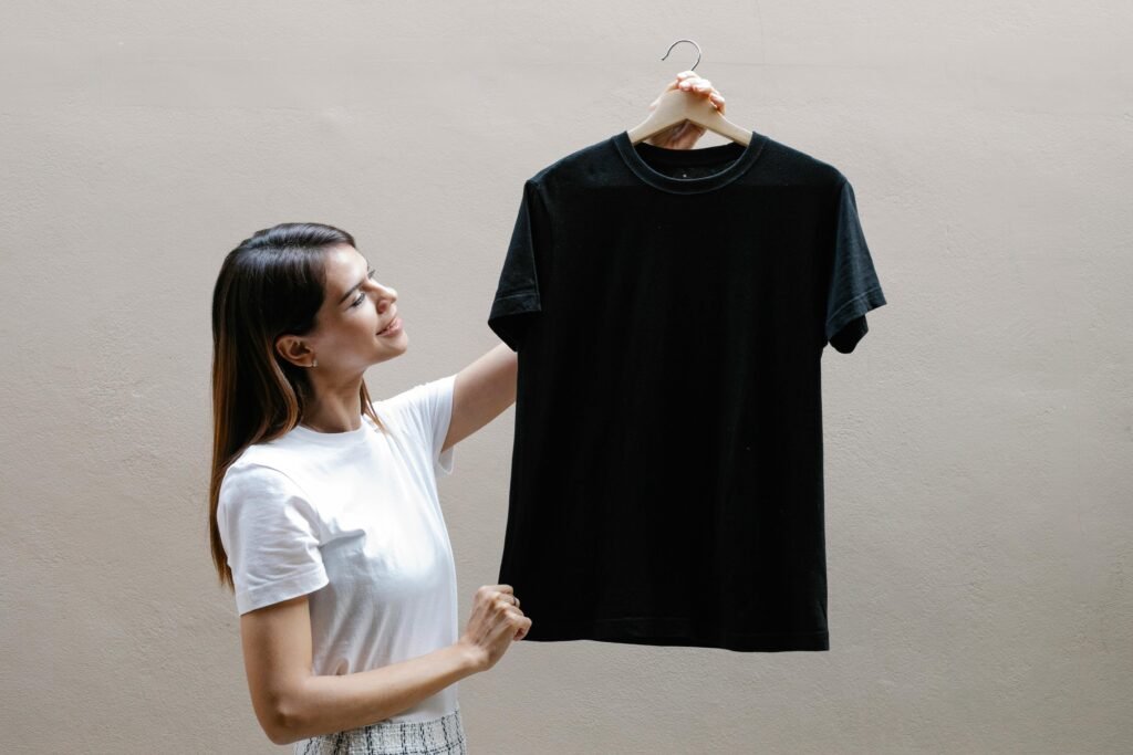 A cheerful woman in a white t-shirt holding a black t-shirt on a hanger against a gray background.