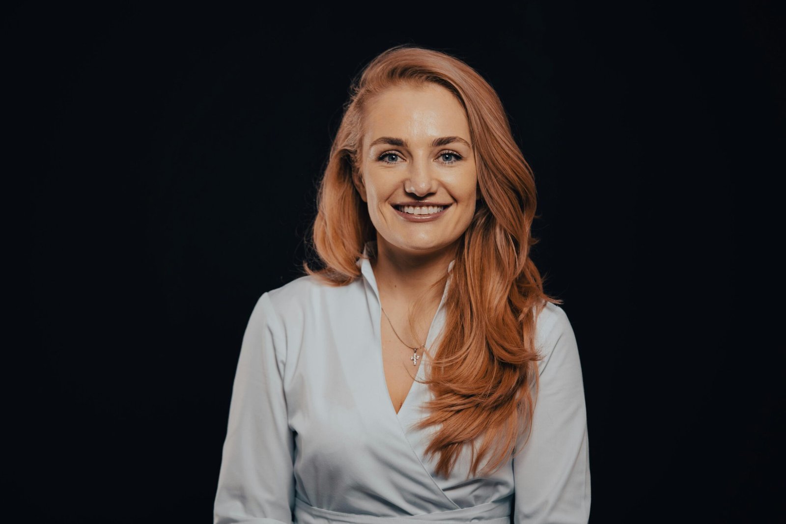 Studio portrait of a smiling woman with red hair in an elegant white dress against a dark background.